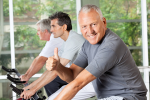 Picture of an elderly man on an exercise bike giving a thumbs up and two men on exercise bikes  next to him as well.