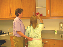 Picture of a Occupational Therapist helping a woman while she reaches for a cup in a cabinet.