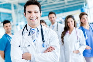 Picture of a male Physician crossing his arms and smiling. There is female and male Physician in the background and two female nurses and one male nurse smiling.